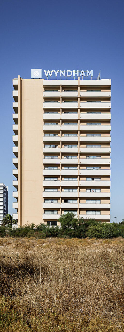 Façade principale d'un immeuble hôtelier Wyndham de plusieurs étages avec balcons alignés, sur fond de ciel bleu, derrière un terrain en friche aux hautes herbes sèches.