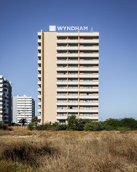 Façade principale d'un immeuble hôtelier Wyndham de plusieurs étages avec balcons alignés, sur fond de ciel bleu, derrière un terrain en friche aux hautes herbes sèches.